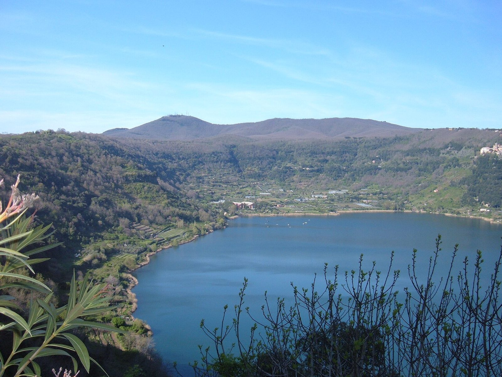 Il lago di Nemi visto dall'alto da Genzano di Roma: l'acqua blu racchiusa in una conca verde di boschi e campi, con i rilievi dei Colli Albani sullo sfondo e il cielo azzurro