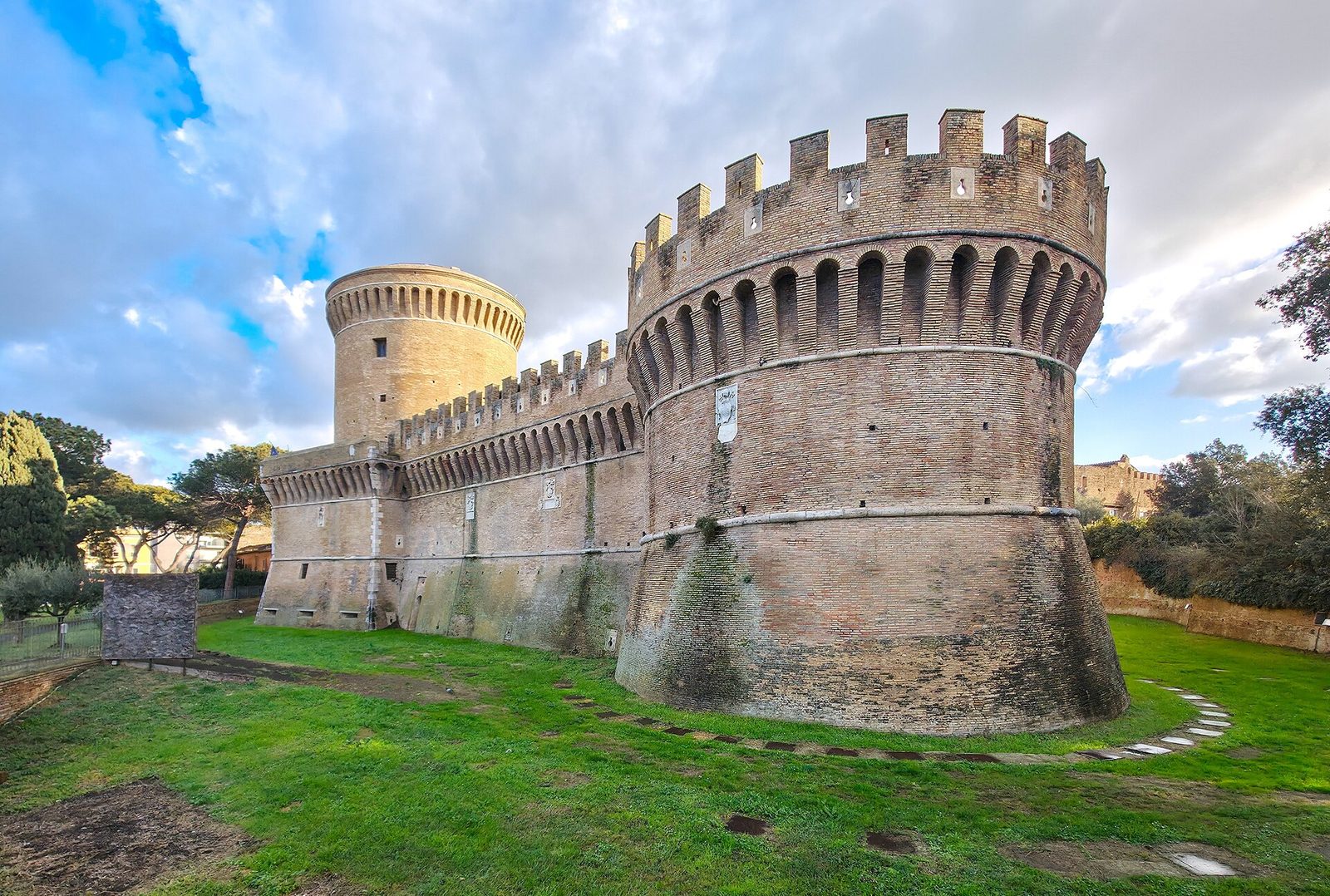Il Castello di Giulio II a Ostia Antica: il grande torrione circolare merlato in mattoni, le mura con beccatelli e il mastio cilindrico sullo sfondo, circondati da un prato verde sotto un cielo nuvoloso