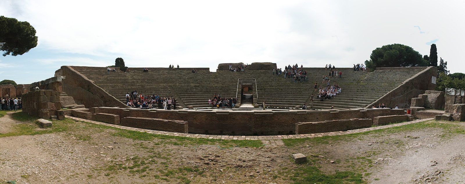 Veduta frontale del teatro romano di Ostia Antica: la cavea semicircolare con le gradinate in pietra, il muro in mattoni dell'orchestra in primo piano e piccoli gruppi di visitatori seduti sui gradini, sotto un cielo velato con pini ai lati