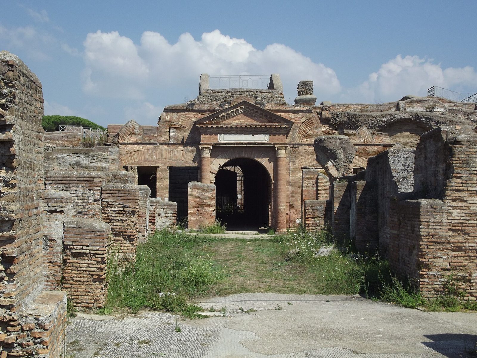 L'ingresso degli Horrea Epagathiana negli scavi di Ostia Antica: un portale in mattoni con frontone triangolare e due colonne ai lati di un arco con cancello, tra muri romani in laterizio e un prato sotto un cielo con nuvole