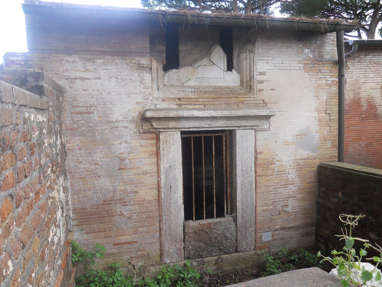 Una tomba a camera in mattoni della Necropoli di Porto all'Isola Sacra: facciata con porta incorniciata in travertino chiusa da una grata e, sopra, una lastra di marmo frammentaria, sotto una copertura moderna con pini sullo sfondo