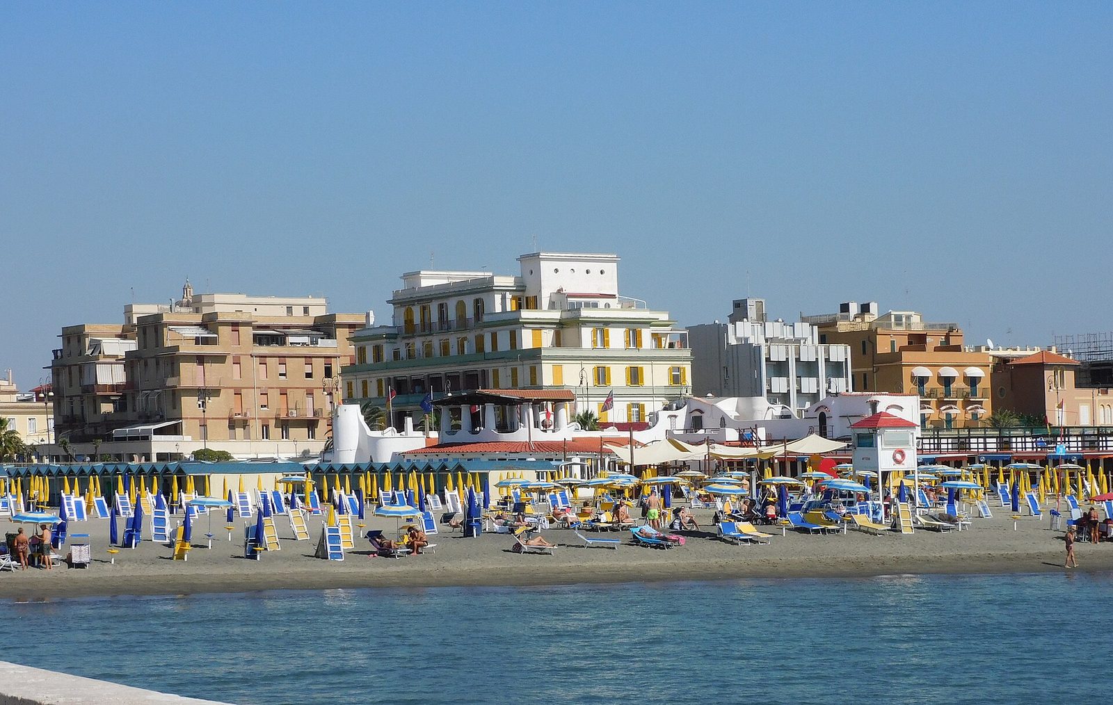 La spiaggia di Lido di Ostia in una giornata di sole, con file di ombrelloni e lettini colorati sulla sabbia e alle spalle i palazzi del lungomare