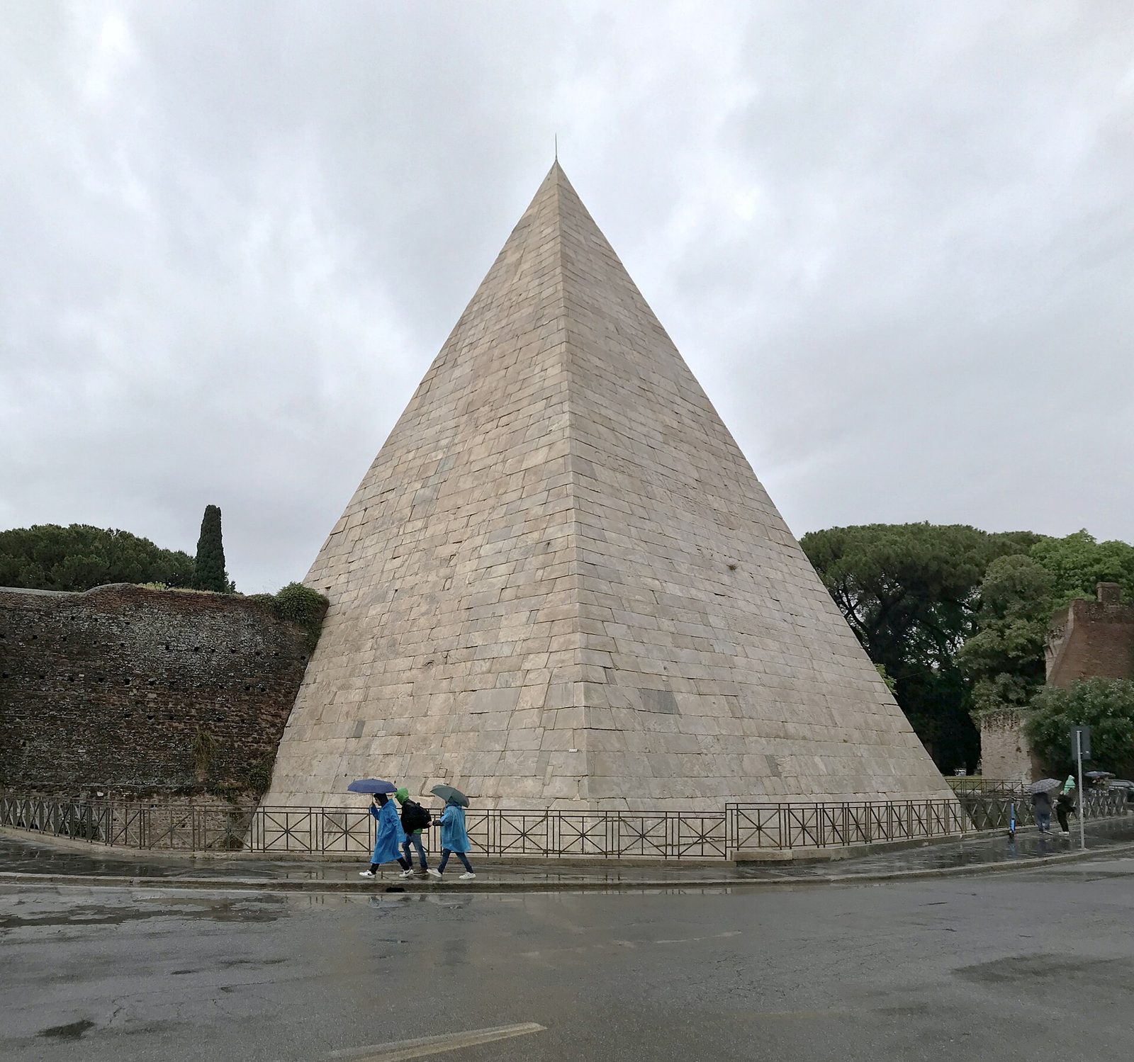 La Piramide Cestia a Roma in una giornata nuvolosa: la grande piramide rivestita di blocchi di marmo chiaro accanto a un tratto delle Mura Aureliane in mattoni, con pini sullo sfondo e alcuni passanti con ombrelli sul marciapiede