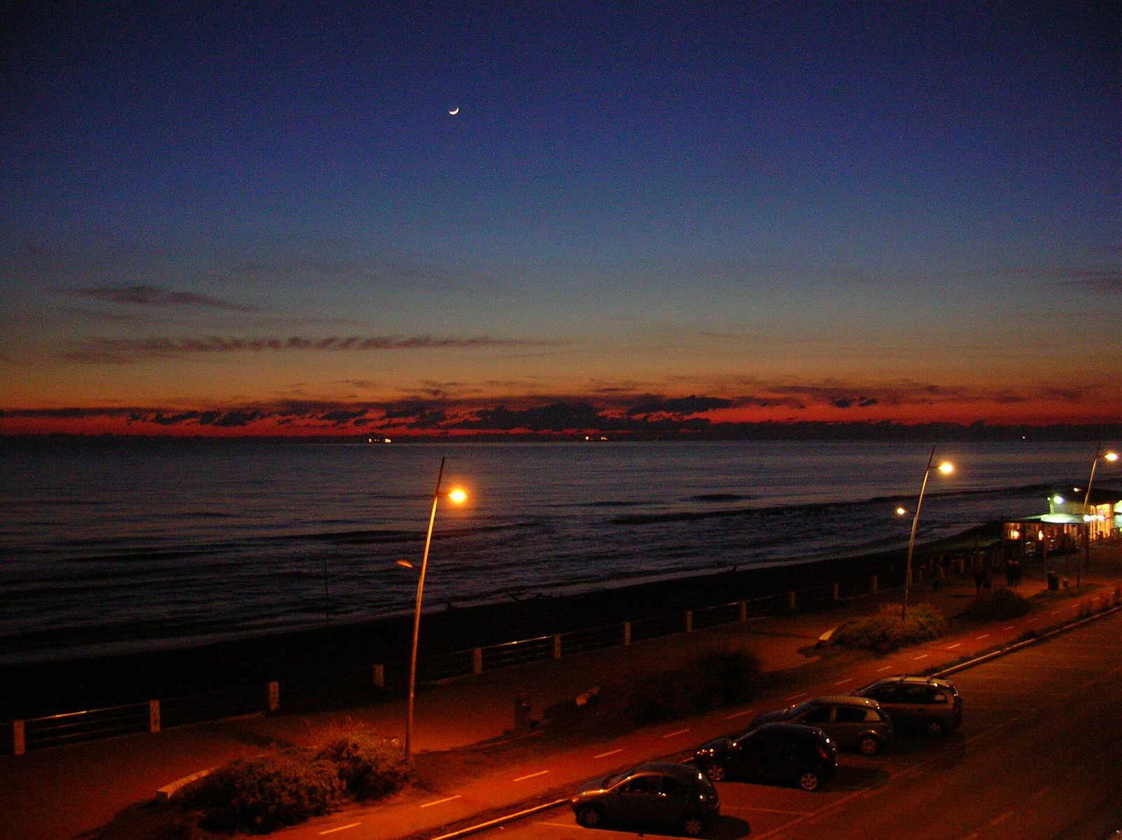 Il lungomare di Ostia al crepuscolo: il mare calmo sotto un cielo blu con una sottile falce di luna e una striscia rossa all'orizzonte, i lampioni accesi, la pista ciclabile e alcune auto parcheggiate lungo la strada