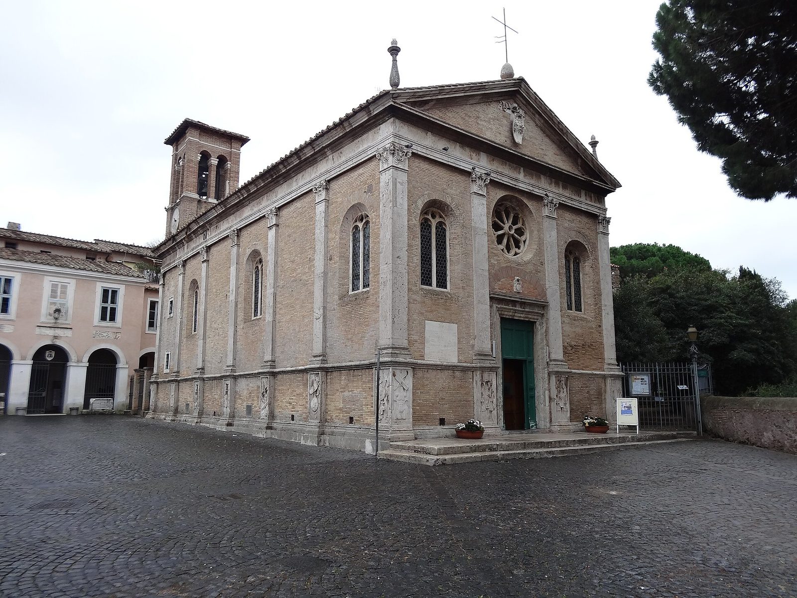 La basilica di Sant'Aurea nel borgo di Ostia Antica: facciata rinascimentale in mattoni con lesene in travertino, rosone e portone verde, campanile a sinistra e piazza lastricata in sampietrini, con un pino sulla destra