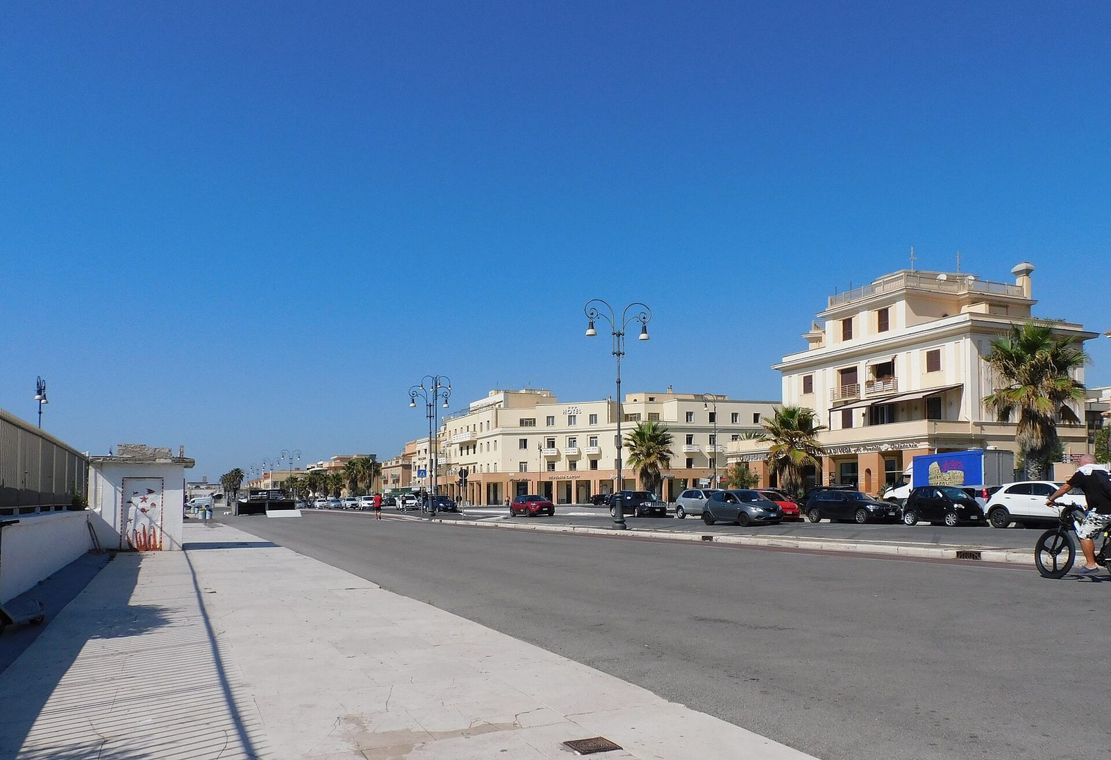 Il lungomare Paolo Toscanelli a Ostia in una giornata di cielo terso: l'ampia carreggiata con lampioni in ghisa, auto parcheggiate e una fila di palazzi chiari del primo Novecento con palme davanti