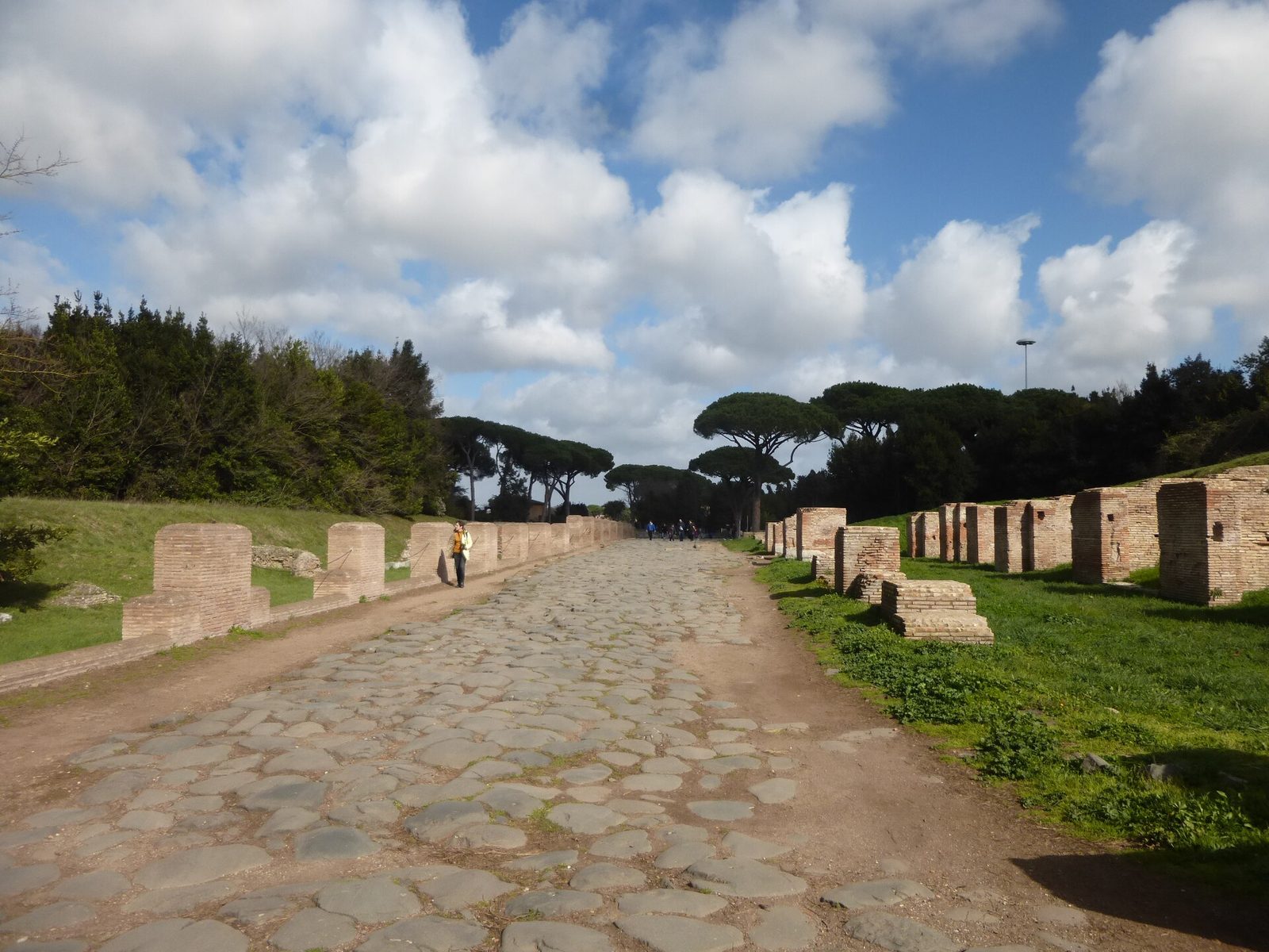 Il Decumano Massimo degli scavi di Ostia Antica: una strada romana lastricata di grandi pietre scure fra ruderi in mattoni e prati verdi, con pini domestici sullo sfondo e qualche visitatore lontano